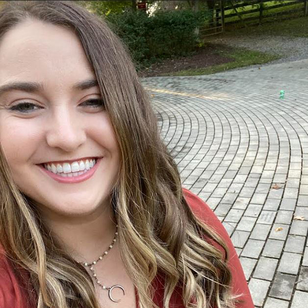 A woman with long, wavy hair smiles at the camera, wearing a red top and a necklace with a crescent moon pendant, against a backdrop of a stone path and greenery.