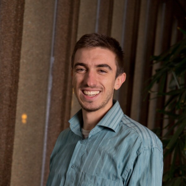 A young man smiling while wearing a light blue striped shirt, standing in front of a textured wall with a plant in the background.