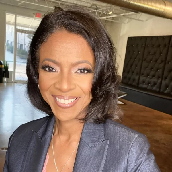 A smiling woman with shoulder-length, slightly wavy hair wearing a dark blazer and a silver necklace, posing in a bright, modern interior with a black tufted sofa in the background.