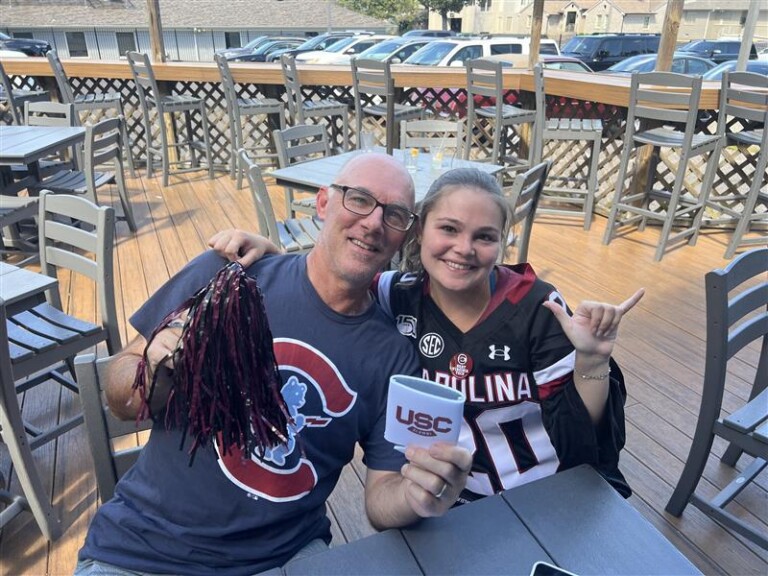 A man with glasses and a dark shirt holding a pom-pom sits next to a young woman in a football jersey, both smiling at the camera. The setting is an outdoor deck with empty tables and chairs visible in the background.