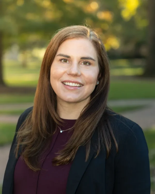 A woman with long brown hair and a friendly smile stands outdoors in a green park setting, wearing a black blazer over a dark maroon top.