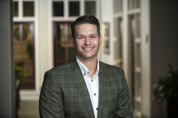 A man in a checked blazer and white shirt smiles warmly, standing in a well-lit indoor setting with a blurred background.