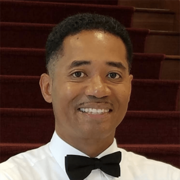 A man smiling while wearing a white dress shirt and a black bow tie, standing in front of a set of wooden stairs with a dark red carpet.