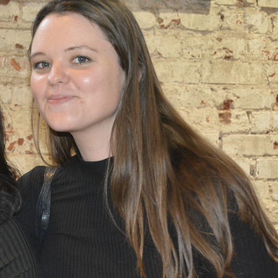 Two women smiling together in front of a textured brick wall; one wearing a black top with long, dark hair and the other with long, light brown hair.