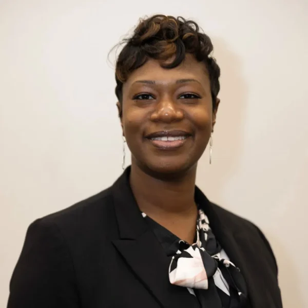 A woman with short, styled hair and earrings smiles confidently at the camera, wearing a black blazer over a blouse with a light-colored, patterned necktie against a neutral background.