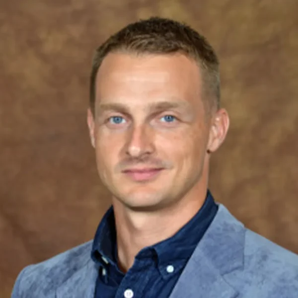 A man with short, light brown hair and blue eyes is smiling at the camera. He is wearing a blue collared shirt under a light blue blazer against a neutral brown background.