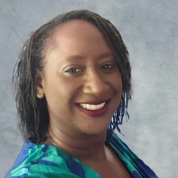 A person with dark, shoulder-length hair styled in loose curls smiles brightly, wearing a patterned blue and green top against a light gray background.
