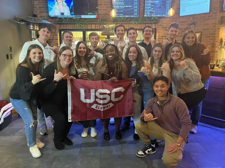 A group of alumni poses together, holding a maroon flag that says 'USC ALUMNI'; the setting is a bar with a casual atmosphere, featuring wooden accents and screens showing sports events. Most of the group members are making hand signs.