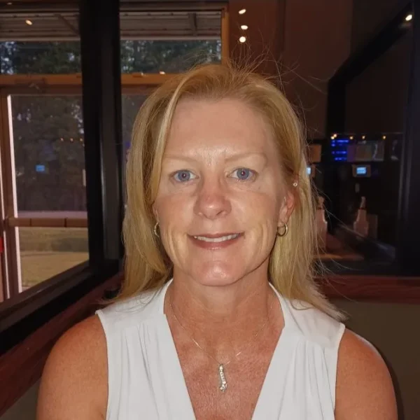 A woman with shoulder-length blonde hair smiles at the camera, wearing a light-colored sleeveless top and a necklace, in a dining setting with blurred background elements.