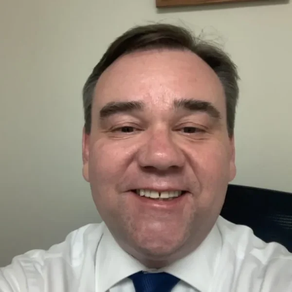 A man smiling in a white dress shirt and blue tie, seated in front of a plain wall, with short brown hair and a friendly expression.