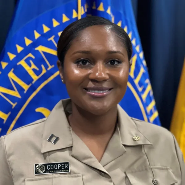 A woman in a tan uniform smiling in front of a blue flag with yellow lettering, displaying the words related to public health.