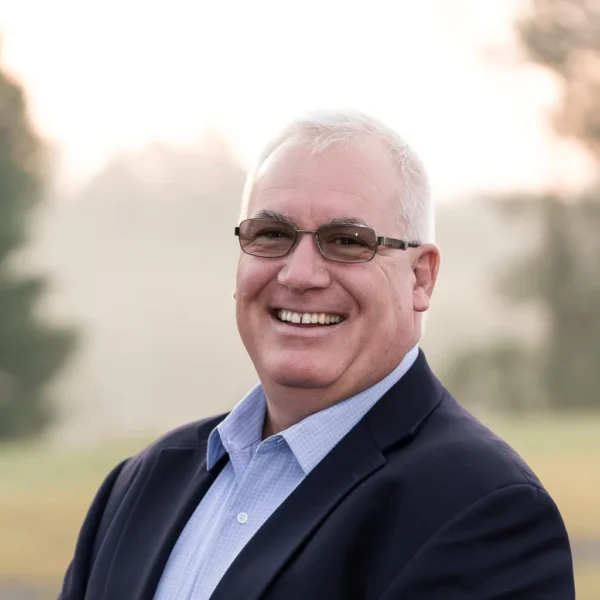 A smiling middle-aged man with glasses, wearing a dark blazer over a light blue shirt, standing outdoors against a blurred background of greenery and soft light.