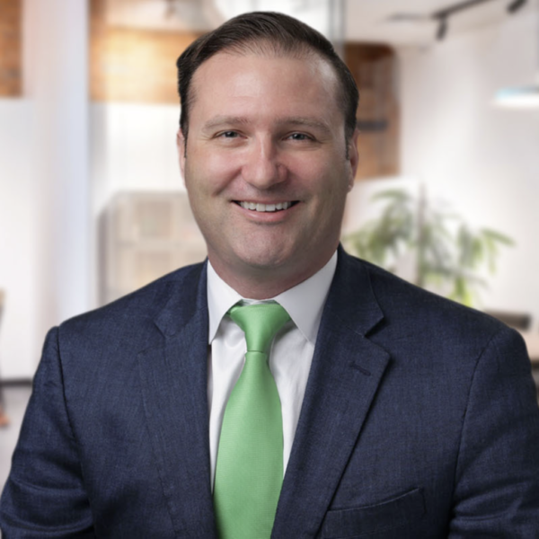 A man with short hair smiles while wearing a blue suit and a bright green tie, set against an office background with plants and soft lighting.