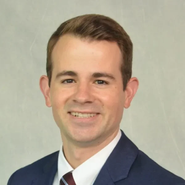 A smiling young man with short brown hair wearing a dark suit and a white shirt with a maroon tie, posing against a light textured background.