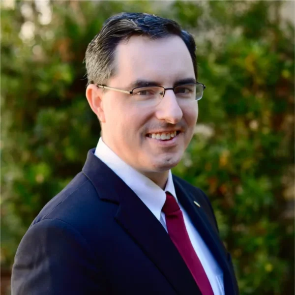 A man smiling and looking to the side, wearing a dark suit and a red tie, with short dark hair and glasses, set against a green foliage background.