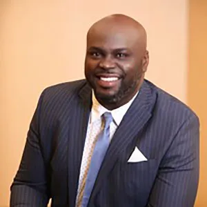 A smiling man with a bald head wearing a navy pinstripe suit and a light blue tie, sitting against a neutral background.