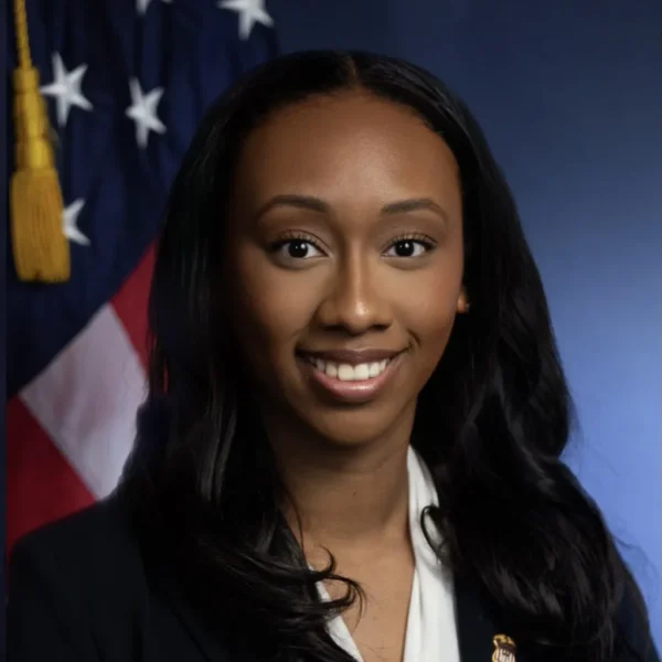 Smiling individual with long dark hair, wearing a black blazer and white shirt, set against a backdrop featuring the American flag and a blue gradient.