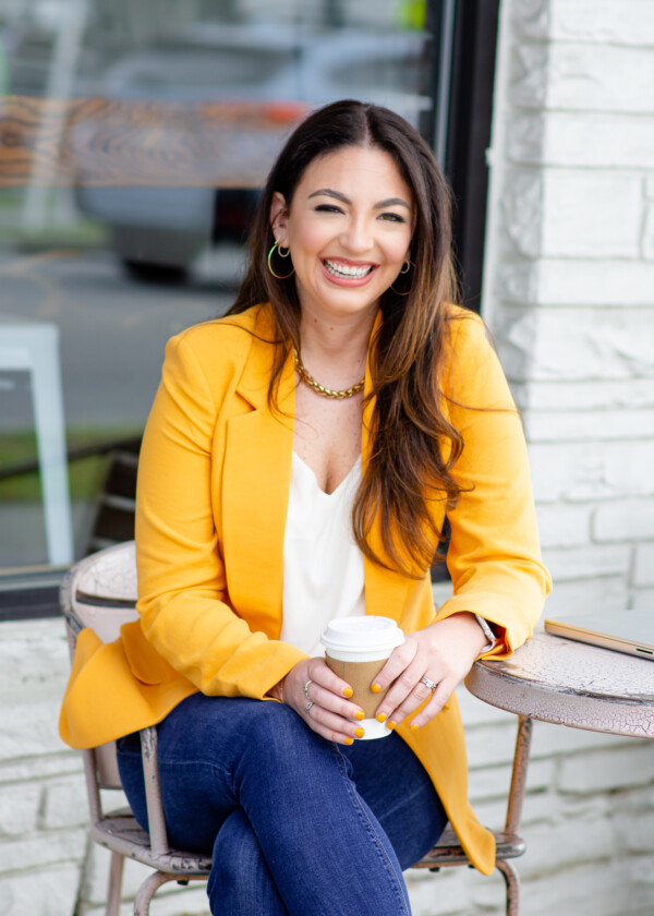 A woman sitting at a small table outside, wearing a bright yellow blazer over a white top. She has long brown hair and is smiling while holding a coffee cup. Her nails are painted yellow, and she is accessorized with hoop earrings and a chain necklace.