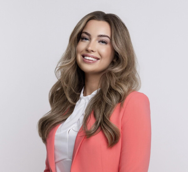 A woman with long, wavy hair smiles confidently while wearing a coral blazer over a white blouse against a light gray background.
