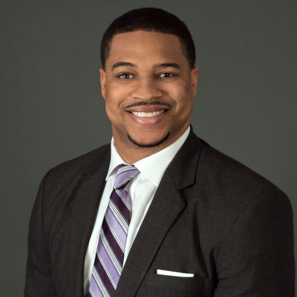 A smiling man in a dark suit with a white dress shirt and a striped purple tie, posed in front of a dark gray background.
