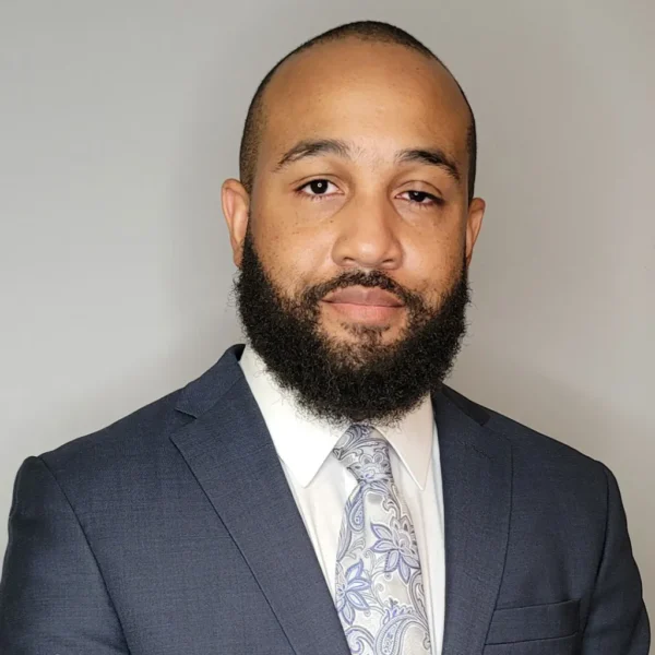 A man with a well-groomed beard and short hair, dressed in a dark suit and a light-colored paisley tie, posing against a plain gray background.