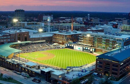 Aerial view of a vibrant baseball stadium filled with fans, with tall buildings and a cityscape in the background during sunset, creating a scenic atmosphere.