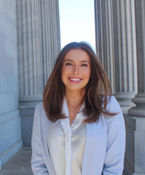 A young woman smiles brightly while standing between two tall, fluted columns. She has long, dark hair and is wearing a light blue blazer over a white blouse.