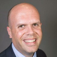A man with a bald head and a wide smile, wearing a dark suit and a light blue shirt, against a neutral gray background.