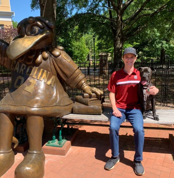 A smiling young man in a red shirt sits on a bench next to a large bronze statue of a bird mascot, holding a black dog on his lap. The setting features trees and a park-like atmosphere.