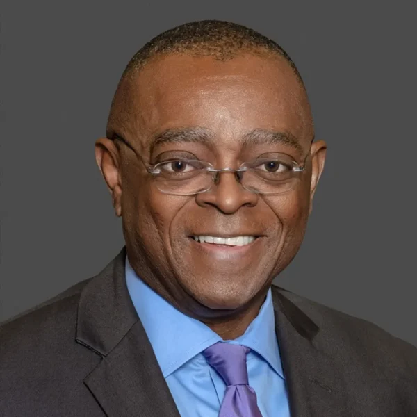 A smiling man with short, closely cropped hair, wearing eyeglasses, a dark suit, and a light blue shirt with a purple tie against a gray background.