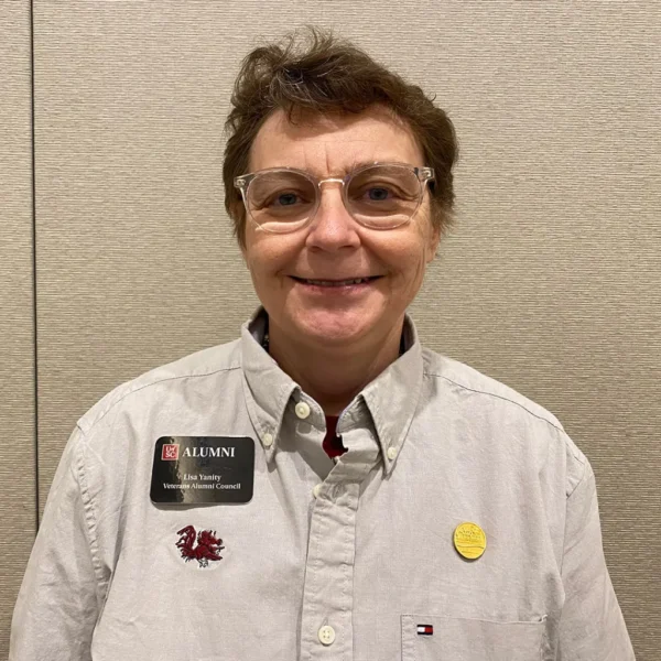 A woman with short brown hair wearing glasses and a light gray button-up shirt, which has a red emblem on the left side, stands in front of a beige wall. She has a name tag that reads 'Lisa Vanity, Alumni Veterans Council' and a yellow button on her shirt.