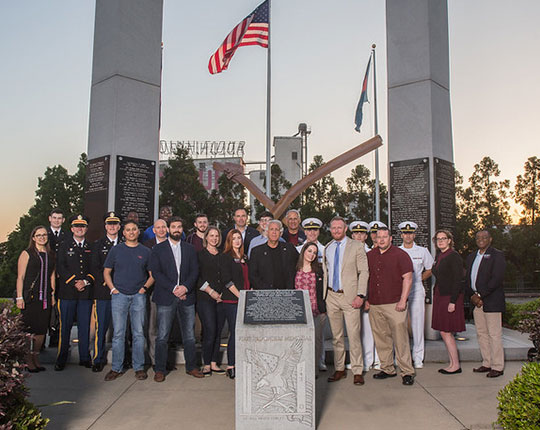 A diverse group of individuals poses together in front of a memorial with tall pillars and flags, surrounded by greenery, at sunset. The group includes military personnel and civilians, standing around a central stone monument with an engraved plaque.