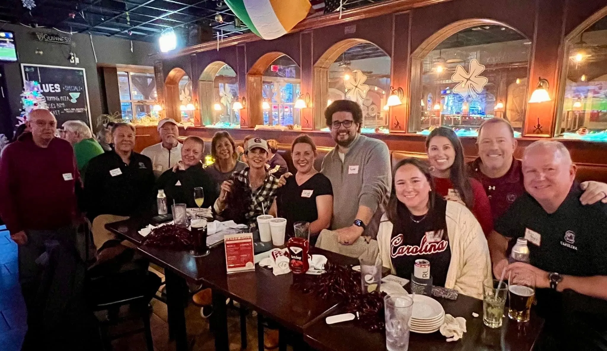 A group of twelve people, including both men and women, are gathered around a table in a lively pub setting, smiling and posing for the photo. The table is cluttered with drinks and plates, and a festive atmosphere is suggested by the decorations in the background, including a Christmas tree and colorful banners. Some individuals are wearing name tags and team apparel.