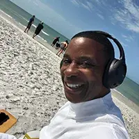 A smiling person wearing headphones sits on a sandy beach, with the ocean in the background and several people walking along the shore.
