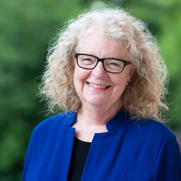 A smiling older woman with curly blonde hair, wearing glasses and a blue jacket, stands outdoors against a blurred green background.