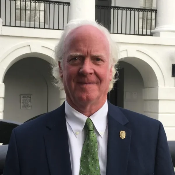 A man with white hair and a light beard poses outdoors, wearing a dark suit and a green tie with a pattern. Behind him, a building with white arches and balconies is visible.