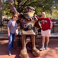 Two people pose next to a large bronze statue of a mascot, wearing a jersey with 'CAROLINA' and the number '04', outdoors with trees in the background.