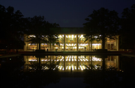 A modern building illuminated at night, reflected in a calm pond, surrounded by trees.