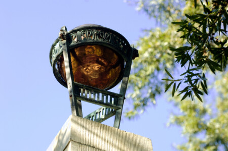 A decorative metal lantern with a copper bowl-shaped top, mounted on a pillar, surrounded by green foliage and a clear blue sky.