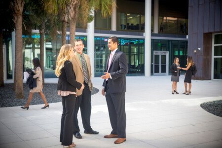 A business setting with three people in discussion: a man in a dark suit and pink tie, a man in a tan suit with a green and purple striped tie, and a woman in a black suit with long blonde hair. In the background, a woman in a beige outfit is walking by, while another pair of women in black suits are conversing nearby under palm trees.