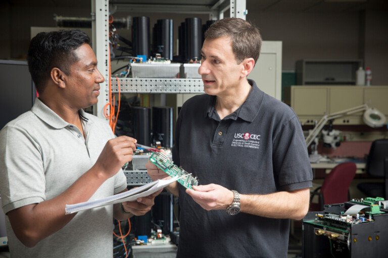 Two men discuss electronics in a lab setting, one holding a circuit board and the other referencing a paper, with equipment and components visible in the background.