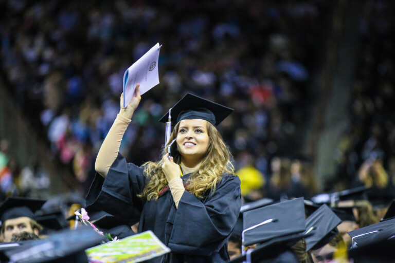 A graduate with long, wavy hair wearing a black cap and gown is joyfully raising a diploma in one hand while smiling at the camera, surrounded by a blurred crowd of fellow graduates during a commencement ceremony.