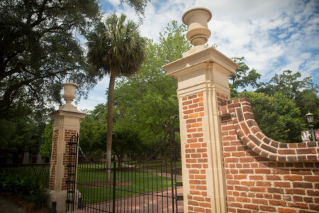 Brick gateposts with decorative urns, surrounded by lush greenery and a palm tree, leading to a manicured lawn under a partly cloudy sky.