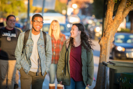 A group of four young adults walking down a sunlit street, with two smiling in the foreground. One is a young man with glasses wearing a denim jacket and gray shirt, while the young woman next to him has curly hair and wears a green jacket. In the background, another woman in a plaid shirt and a man in a black 'Carolina' sweatshirt are also walking.