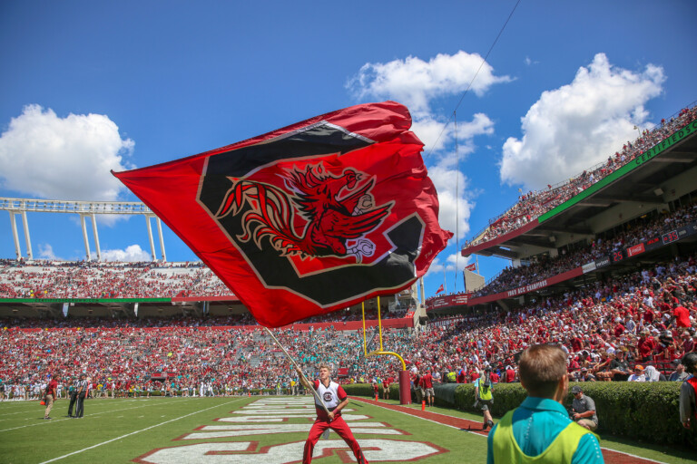 A cheerleader waves a large red flag with a Gamecock emblem at a football game, surrounded by a cheering crowd in a stadium under a blue sky with scattered clouds.