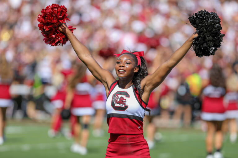 A cheerleader wearing a red and white outfit and holding red and black pom-poms raises her arms in excitement on a football field, with a crowd of fans in the background.