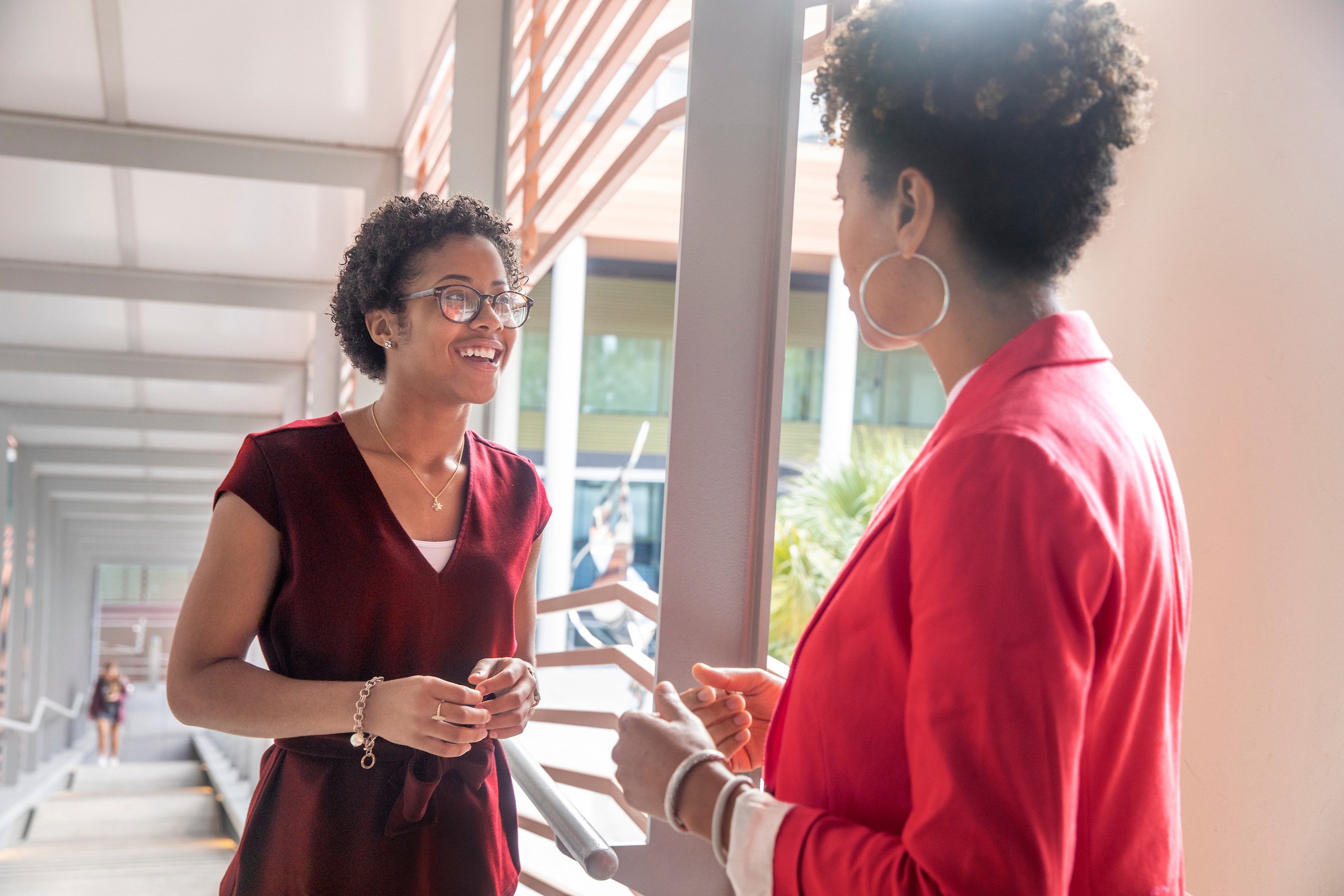 Two women are engaged in conversation on a staircase with a modern design; one is wearing a burgundy top and glasses, smiling while the other wears a red blazer.