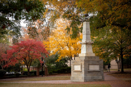 A stone monument with a tall, octagonal structure is situated in a park, surrounded by vibrant autumn foliage in shades of red, orange, and green; a person is walking in the background along a brick path.
