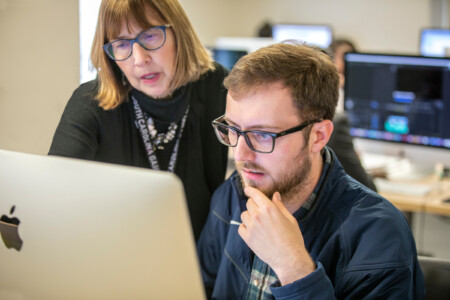 A woman with shoulder-length hair wearing glasses and a black top leans over a young man sitting at a computer, looking intently at the screen while he rests his chin on his hand, both appearing engaged in a discussion.