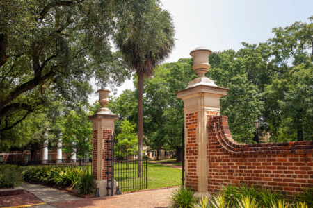 An ornate brick entrance gate with stone pillars featuring decorative urns, surrounded by lush greenery and trees, leading to a landscaped area.